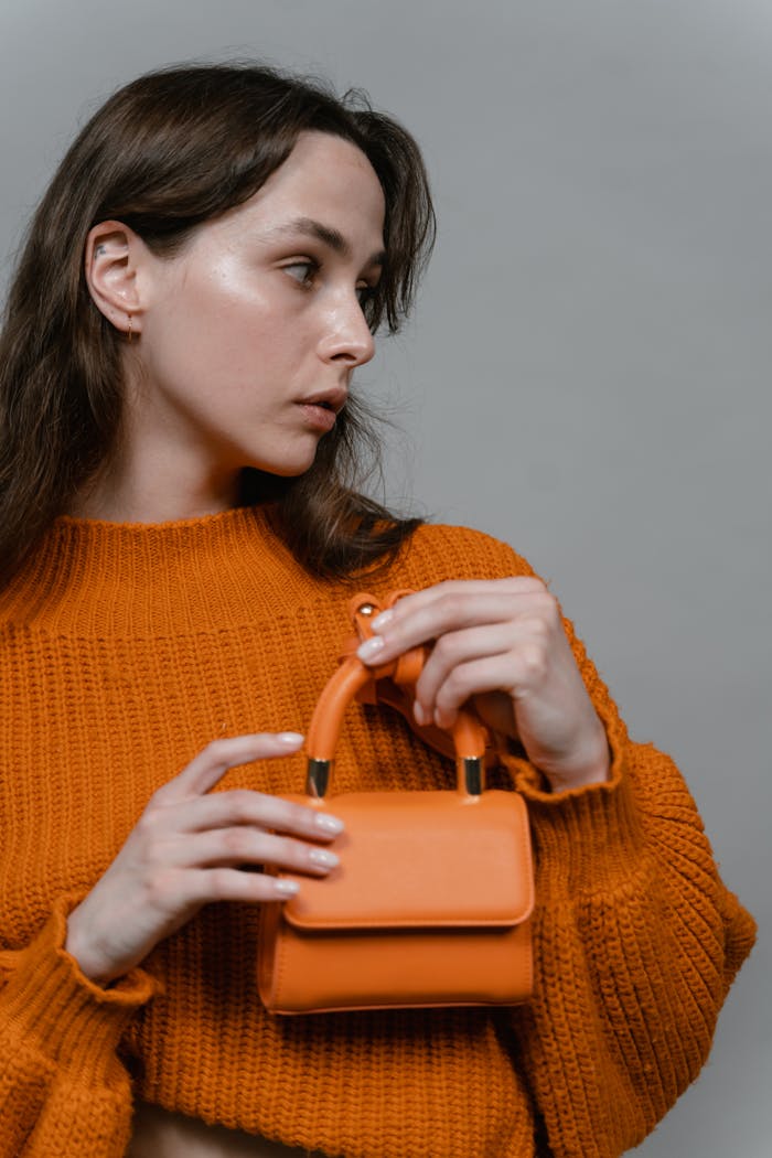 Stylish woman in orange sweater holding a matching handbag against a neutral background.